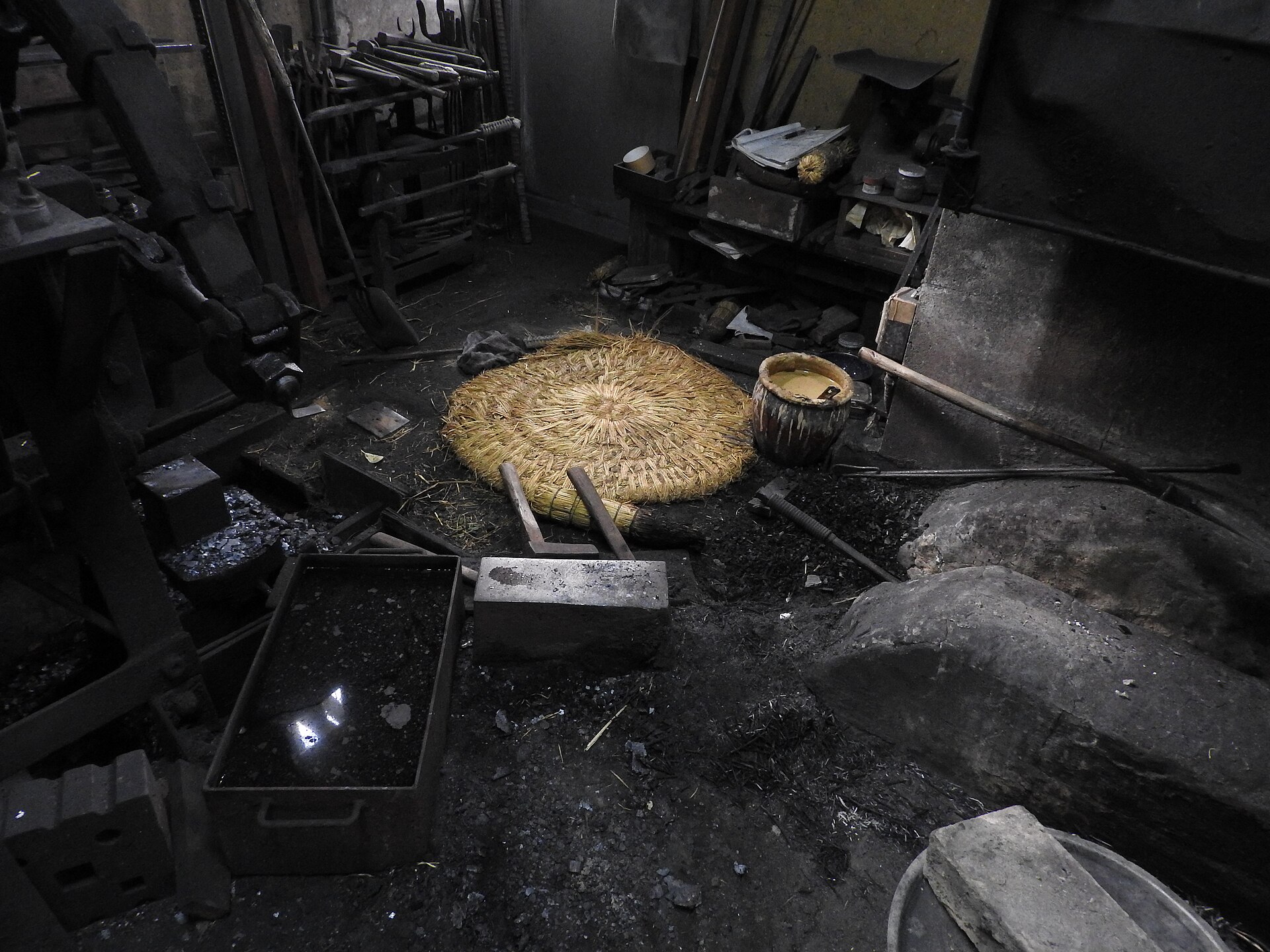 A dark Japanese forge: hammers on an anvil, a straw mat and a clay pot of quench water on a soot-black floor