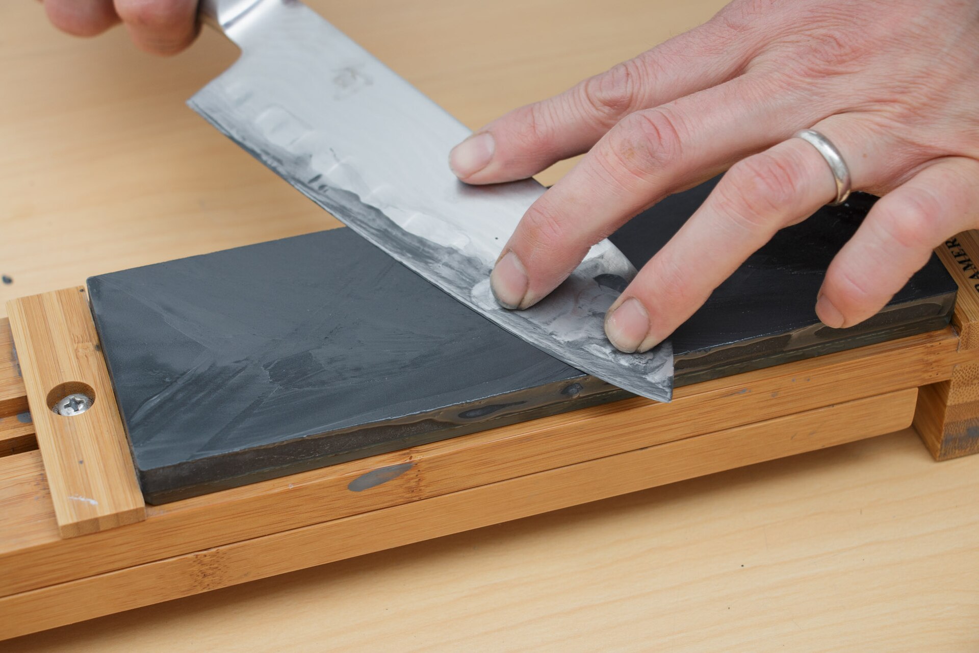 Two hands holding a kitchen knife flat against a dark Japanese whetstone on a bamboo base